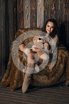 Cute girl in knee socks and sweater with Teddy bear in her hands sitting in armchair in wooden room