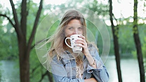 Cute girl with long hair holds a cup of tea or coffee in hands against the blurred nature background. Pretty woman in