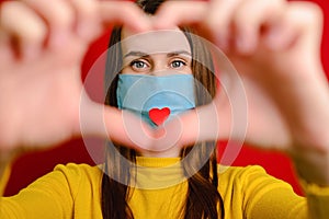 Cute girl making heart shape hand gesture looking at camera, wear medical mask with a red heart