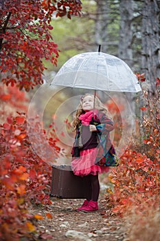 Cute girl with an old suitcase and an umbrella in her hand on a footpath in the autumn forest