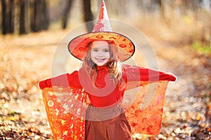 cute girl in an orange costume and a witch hat smiles on the background of the forest, celebrating Halloween