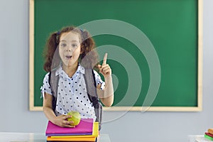 Cute girl with pile of books and apple pointing up near chalkboard at classroom. Little child having idea at school