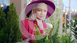 Cute girl in pink hat spraying plants in indoor garden orangery. Florists girl spraying green flowers and tree for care
