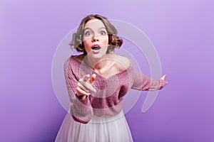 Cute girl with pink makeup expressing amazement and pointing finger. Indoor photo of curly shocked woman isolated on