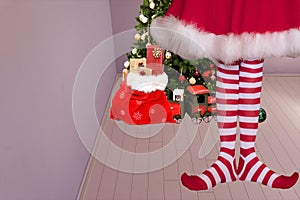 Cute girl with pointed elven feet wearing elf legging standing in a room with a christmas tree and santa claus bag full of