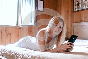 Cute girl portrait. woman lies on the bed and uses a smartphone. Wood interior decoration, bright colors, soft light from the