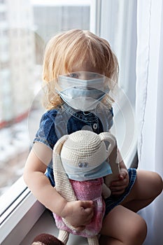 Cute girl preschool child sits on the window in a medical mask and hugs a toy. new normality family