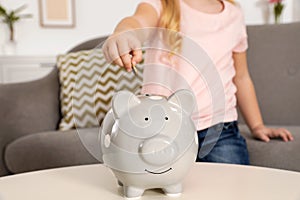 Cute girl putting coin into piggy bank at table. Saving money