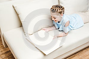 Cute girl reading book while lying on sofa at home