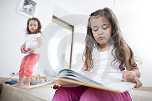 Cute girl reading book with sister playing in background at home