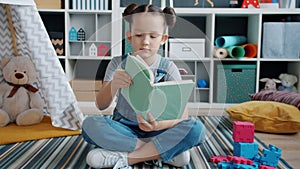 Cute girl reading book sitting on floor in nursery room concentrated on activity