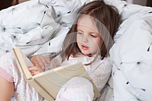 Cute girl reading her favorite fairy tale, child sitting in bed with book, Kid preparing to go to bed, spending pleasant time in