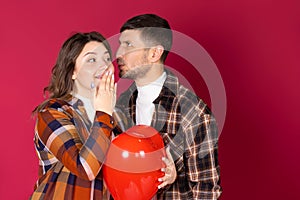 Cute girl with a red heart-shaped balloon and a man whispering something to her. Red background and side space.