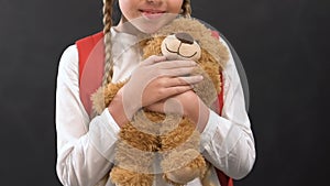 Cute girl with rucksack hugging teddy bear, happy childhood, primary school