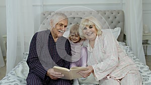 Cute girl with senior retired grandmother and grandfather sitting on bed reading book in bedroom