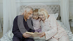 Cute girl with senior retired grandmother and grandfather sitting on bed reading book in bedroom