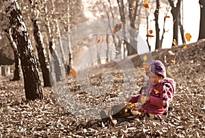 Cute girl sitting on fallen autumn leaves while leafs falling and playing with dolls