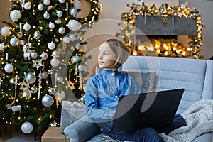 A cute girl is sitting on a sofa with a laptop in a decorated room for Christmas. Communication, meetings and training online. He