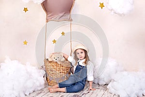 Cute girl sitting in the studio on the background of a balloon, stars and clouds and holding a teddy bear. The little girl is drea