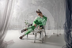 Cute girl smiles and drinks coffee with cupcakes in studio. Young woman in green suit poses on gray background