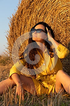 Cute girl in sunglasses sitting near straw roll on field.