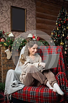 A cute girl in a sweater is sitting in a chair near the Christmas tree and looking at a laptop