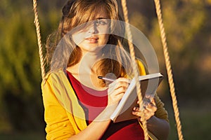 Cute girl on a swing with notebook and pen keeps a diary of feelings in nature, a woman composes and writes, romantic mood photo