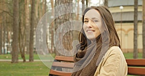 Cute girl talking to camera and smiling sitting in autumn park on bench and starts to type on the phone.