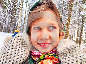 Cute girl in a traditional Russian headscarf and mittens on winter forest. Closeup portrait of a child in folk clothes