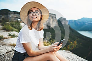 Cute girl traveler in hat and glasses resting during their vacation in nature, female tourist uses mobile phone while enjoying