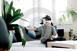 Cute girl tries to touch something that is displayed in goggles of virtual reality, sitting on the sofa