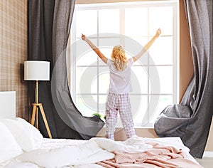 Cute girl wakes up in her bedroom and opens   curtains in the morning