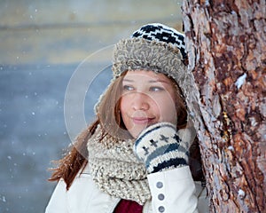 Cute Girl in warm winter clothes smiles flirtatiously during snowfall in front of a wooden barn with lots of snow falling from the