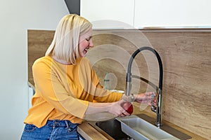 Cute girl washing apple in kitchen sink. Young Woman cleaning her fruit with water under the tap. Daily intake of vitamins with