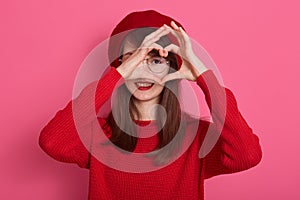 Cute girl wearing beret, sweater and spectacles, showing heart gesture with her fingeres in front of her face to her boyfriend,