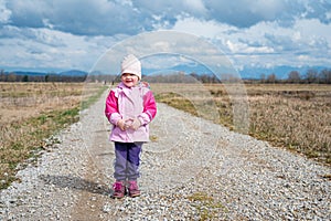 Cute girl wearing pink jacket standing on road running through fields.