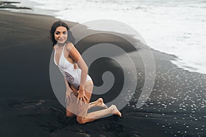 Cute girl in white bathing suit posing on black sandy beach. model resting near ocean