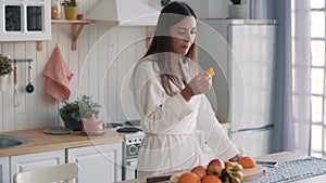 Cute girl in white bathrobe eats slice of orange in kitchen, slow motion