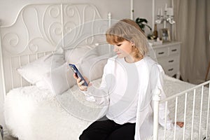 A cute girl in a white shirt and with a phone is lying on the bed