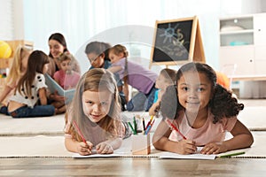 Cute girls drawing on floor while kindergarten teacher reading book to other children