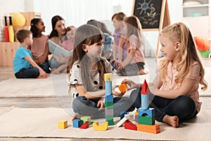 Cute girls playing with building blocks on floor while kindergarten teacher reading book to other children