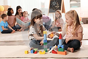 Cute girls playing with building blocks on floor while kindergarten teacher reading book to other children