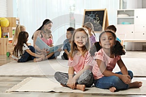 Cute girls sitting on floor while kindergarten teacher reading book to other children