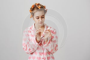 Cute and glamorous woman in hair curlers and pyjamas with hearts, painting nails while being over gray background. Girl