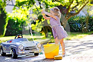 Cute gorgeous toddler girl washing big old toy car in summer garden, outdoors. Happy healthy little child cleaning car