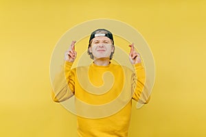 Cute guy in a cap caresses the desire by crossing his fingers on a yellow background and smiling, isolated on a yellow background