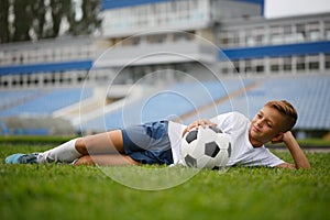 A cute guy with a soccer ball laying on a green grass and on a stadium background. A football player in the outdoors.