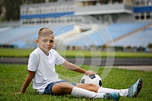 A cute guy with a soccer ball sitting on a green grass and on a stadium background. A football player in the outdoors.