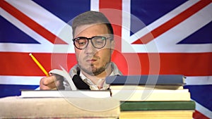 Cute guy student with glasses fluently leafs through a book from a pile of books on the background of the flag of Great