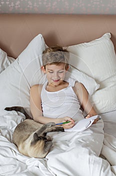 Cute handsome blonde boy lying in white bed and drawing. Boy smiling and playing with cat. Relax time